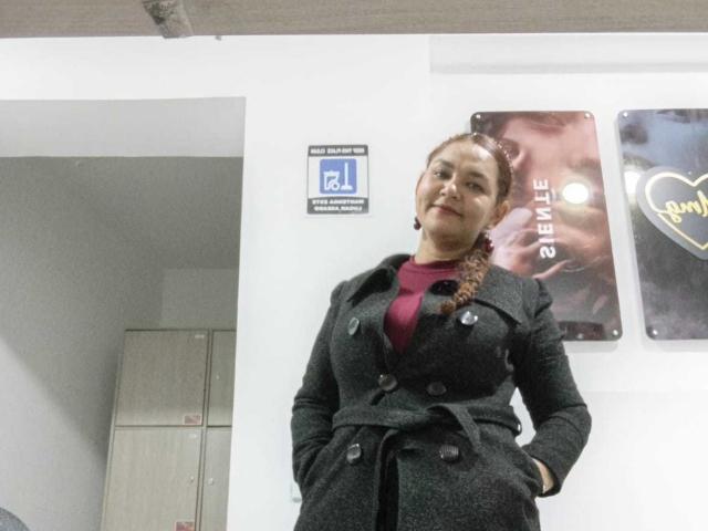 VivianBig with braided hair, dark green coat, red shirt, standing, hands in pockets, slight smile, white wall, locker in background, partial poster on right.
