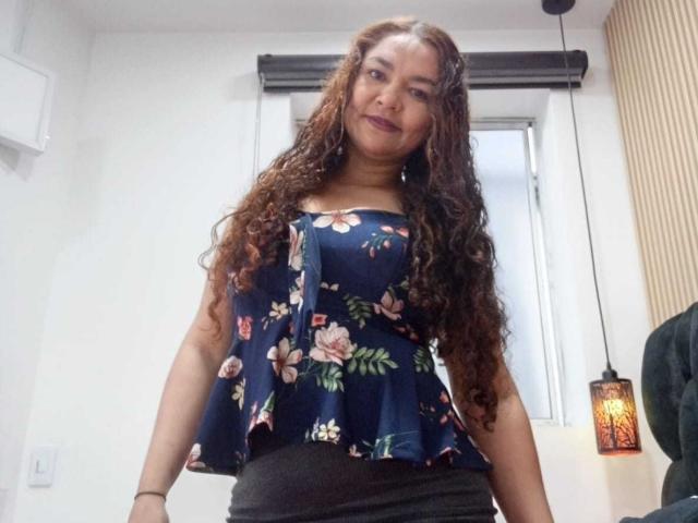VivianBig with long curly brown hair, wearing a blue floral top, showing her small tits, black skirt, smiling, in a room with a hanging lantern.