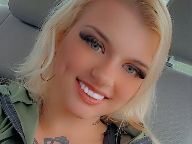 Blonde BarbDollas with green eyes, full white teeth, and long eyelashes, smiling in a car, wearing a green hoodie, has a tattoo on her chest, and hoop earrings.