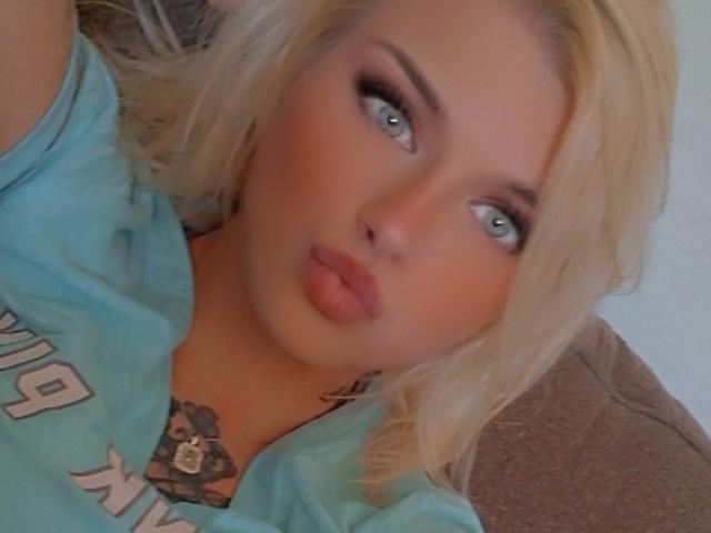 Blonde BarbDollas with pouty lips, blue eyes, and a tattoo near her tits, wearing a light blue shirt, lying on a brown couch. Her makeup is smoky.