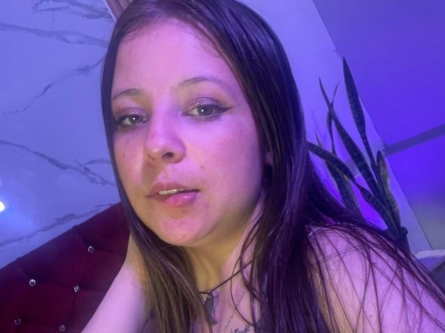 Close-up photo of GraceRedMiller. She has long, straight brown hair, light skin, and subtle freckles. Her eyes are slightly downcast with light green eyeshadow, and she's looking at the camera with a slight, neutral expression. She's wearing a black necklace with a small pendant. The background has a purple-blue light, and a tufted red velvet surface is visible on the left. A potted plant is in the right background. The image has a soft, purple-blue light cast on her face.