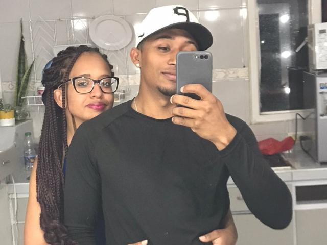 LilithAndAsmodeo with braids, glasses, and pink lipstick, hugs a dude in a black shirt and white cap, holding a phone for a selfie. His dick is not visible.
