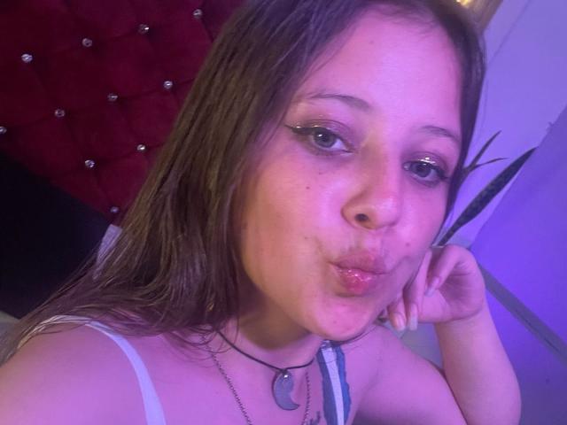 Close-up photo of GraceRedMiller with light brown hair, wearing a white spaghetti strap top, and a dog tag necklace. She has green eyes, winged eyeliner, and is puckering her lips in a pouty expression. Her right hand is resting on her cheek. The purple and red background includes a tufted red headboard and a white wall. The image has a soft, purple-tinted light, emphasizing her facial features and the texture of her hair. Her skin is light with some freckles.