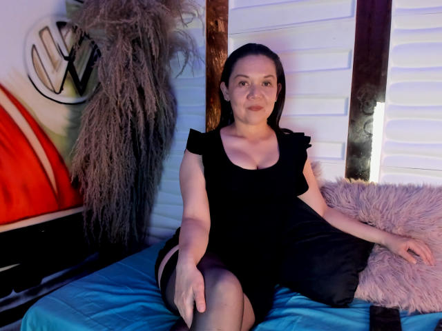 MiilaToro with small tits, wearing a black dress and sheer black stockings, sits on a blue bed with a fluffy pink pillow, and a black pillow. She has dark hair.