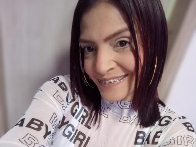 KatteRouse, com um sorriso adorável, dentes de metal brilhantes e um corte de cabelo bob preto, vestindo uma blusa branca estampada e usando brincos de argola dourados, olhando para a câmera..