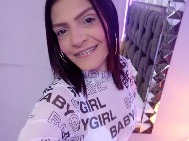 KatteRouse, con capelli castano scuro, indossa una maglietta bianca trasparente con la scritta "BABY GIRL", sorride mostrando l'apparecchio ortodontico, grandi orecchini a cerchio e luci viola sullo sfondo..