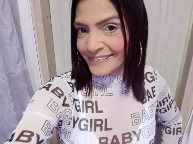 KatteRouse has medium-length black hair, wearing a white long-sleeve shirt with "BABY GIRL" in black letters, smiling with glittery teeth, and large gold hoop earrings.