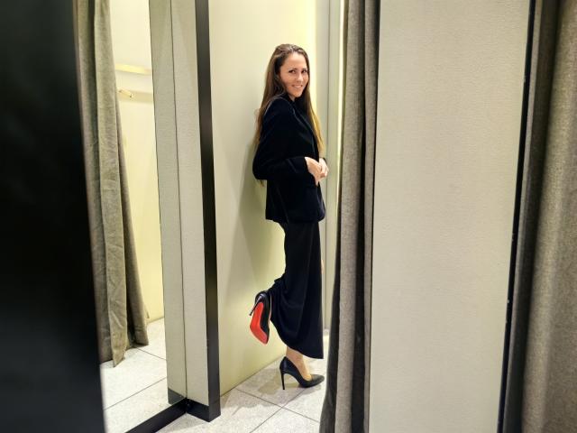 MissKinney stands in a store's changing room, facing a wall with her back partially turned. She has long brown hair and wears a black velvet jacket and black trousers. She holds the jacket's sleeve with her right hand. Her black high heels have red soles. The room has beige walls and grey curtains. The floor is tiled. The composition focuses on her from the side, highlighting her relaxed posture and the contrasting textures of velvet and trousers.