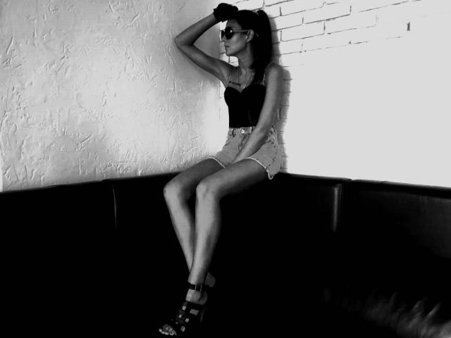 MissKinney with dark tits in a black top, wearing high-waisted shorts and strappy heels, sitting on a black couch, hand in hair, against a white brick wall.