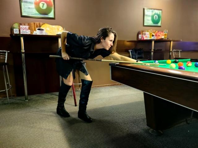 MissKinney with brown hair, black leather dress, and knee-high boots, bending over a pool table, holding a cue stick, aiming at colorful balls. Her ass is prominent.