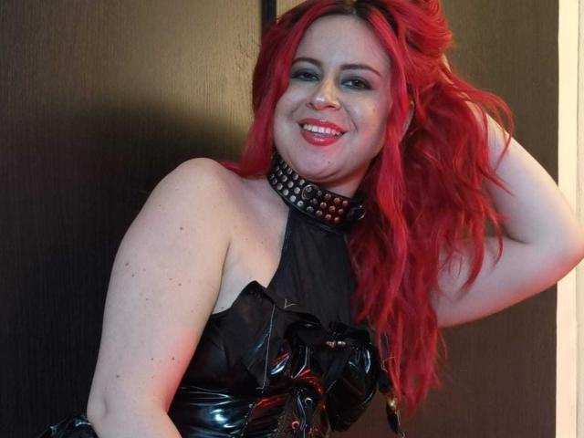 NatySilvy with red hair, smiling, wearing a black leather halter top with spiked choker, hand in hair, showing pale skin and small tits, in a photo.