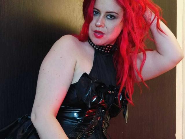 NatySilvy, con un cabello rojo brillante, vestida con un vestido de cuero negro con paneles brillantes y reflectantes, y un collar con púas, apoyada contra una pared oscura, con la mano en el cabello, luciendo seductora..