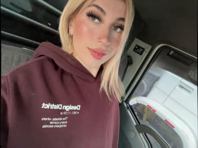 SammyViolet is a blonde woman with light skin and full makeup, including long lashes and pink lipstick. She's wearing a maroon hoodie with white text. Her head is slightly tilted, and she's looking to the side with a subtle smile. The image has a casual selfie style, with soft, natural light. Her makeup is precise, with a slight blush on her cheeks. The composition is tight, focusing on her upper body and face.