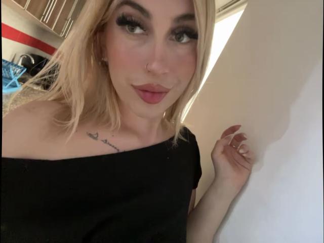 SammyViolet has blonde hair, dark eyebrows, and thick, black eyelashes. She wears a black off-the-shoulder top, exposing a small tattoo on her left collarbone that reads "love" in cursive. She has a nose ring in her right nostril and a subtle smile with pink lipstick. Her right hand touches a white wall. The image is a close-up selfie. The composition focuses on her face and upper chest.