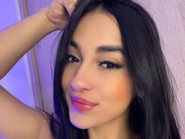 Close-up of EmmaWalter. She has long, straight black hair, and dark, thick eyebrows. Her brown eyes are slightly upturned, with long, dark eyelashes. She's smiling subtly with pink, glossy lips. Her right arm is raised, resting on her head. Her skin is smooth and slightly flushed. The lighting is blue, highlighting her cheek. The texture of her hair is shiny and sleek. Her expression is calm and confident.