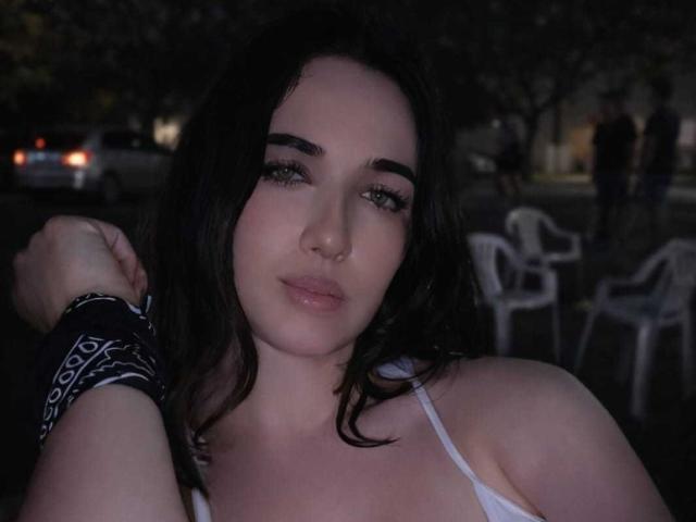 Close-up of LorensStar with pale skin and dark wavy hair. She has a small nose ring in her right nostril and wears a white spaghetti strap top. Her eyebrows are thick and dark, with subtle eyeliner and mascara. She has a relaxed, slightly smiling expression. Her right arm, wearing a black and white patterned wristband, is raised. The background is dark and blurry, with indistinct objects. The image has a soft, low-light effect.