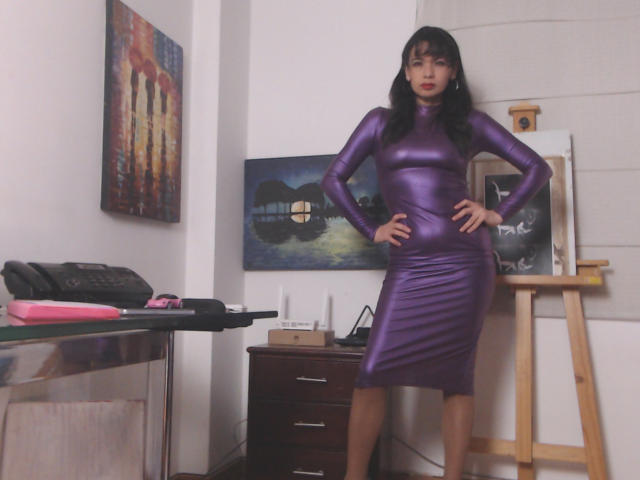 AfroditaRgenis stands in a room, wearing a tight, shiny purple dress that hugs her curves. She has black hair with bangs, red lipstick, and stands with hands on hips. Behind her, a wooden easel holds a partially finished painting. Another painting on the wall shows a nighttime scene with a bridge. A desk to the left has art supplies, including a black bag and pink cloth. The room is well-lit, highlighting the purple dress and AfroditaRgenis's confident stance.