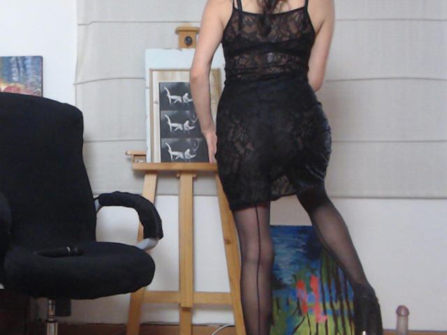 AfroditaRgenis stands with her back to the viewer, wearing a black lace dress and sheer black stockings. Her dress is form-fitting, highlighting her slim build and partially exposing her buttocks. She has dark, wavy hair. In front of her, a wooden easel holds a blackboard with white "bee" letters. A black chair is on the left, and a colorful painting is on the floor. The room has a beige curtain.