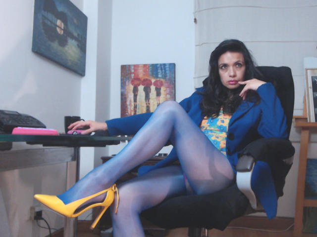 AfroditaRgenis, with blue sheer pantyhose and yellow high heels, sits in a black chair, legs crossed, wearing a blue jacket and yellow patterned top, looking seductively at camera.