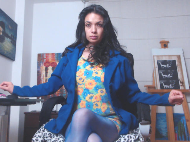 AfroditaRgenis, con cabello largo y negro, chaqueta azul, vestido floral amarillo, medias azules, sentada en una silla de estampado de leopardo, con los brazos extendidos, mirando a la cámara, en una habitación acogedora..