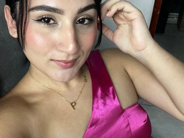 HiilaryDolce with pink satin top, showing her tits, has fair skin, black hair, and a gold necklace, smiling with a hand near her head, looking cute and sexy.