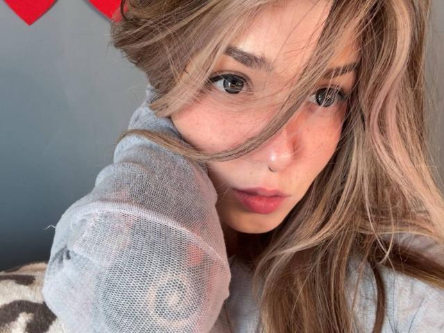 IrishReversed has long, messy blonde hair, gray eyes, and full pink lips. She's wearing a sheer gray shirt, with her hand resting on her head. Her nose is small.