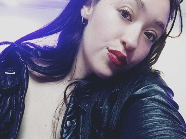 Alanna Scott mit nassen, schwarzen Haaren, die eine schwarze Lederjacke, roten Lippenstift und Hula-Ohrringe trägt, lächelt leicht, wobei ihre Brüste zu sehen sind, in einem Selfie mit blauem Licht..