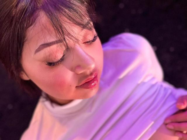 AmeliePiere has light skin with pinkish blush on her cheeks. She has dark brown hair with bangs partially covering her forehead. Her eyes are closed with long, dark eyelashes and pink glossy lips. She wears a white, ribbed top that's slightly shiny under purple and pink lighting. Her right hand is on her chest. The image focuses on her face and upper torso.