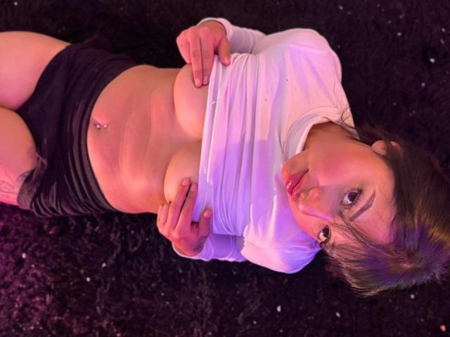 AmeliePiere lies on a black, textured carpet. She has light skin, brown hair, and dark eyebrows. She wears a white long-sleeve shirt, pulled up to expose her large, round breasts with a nipple piercing. She has black shorts and a small tattoo on her right thigh. Her head is tilted slightly, and she looks directly at the camera with a subtle, slightly parted-lip expression. Soft, purple lighting highlights her upper body.