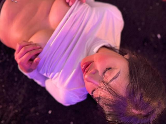 AmeliePiere lies on a dark surface, eyes closed, with a slight smile. She has light skin, brown hair, and is wearing a white shirt pulled up to expose her large breasts and a navel piercing. Soft purple and pink lighting highlights her upper body. Her lips are glossy and red. The image has a sensual, intimate feel with a focus on her exposed chest and relaxed expression. The texture of the shirt is smooth, contrasting with the dark background.