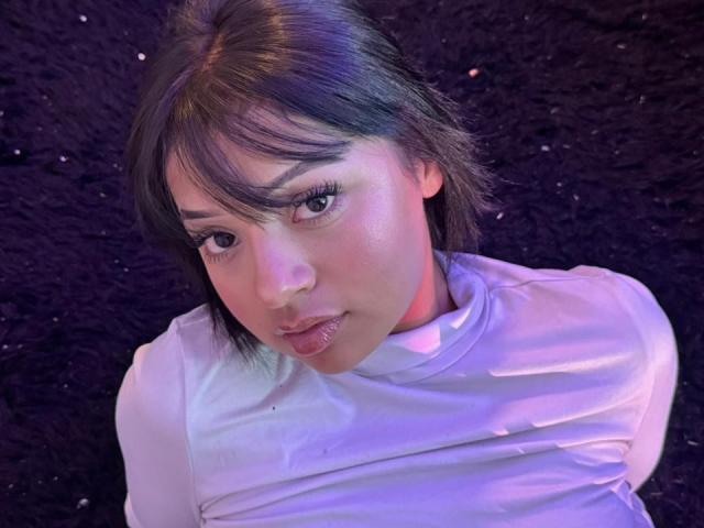 AmeliePiere has short, dark brown hair with slightly wavy strands. She has light skin with a subtle pinkish tint. Her lips are pouted, and she looks up with slightly parted, glossy lips. She wears a shiny, white long-sleeve top that reflects light. The image has a purple-blue light cast on her face and upper body. Her eyes are slightly downward, giving a somewhat innocent or curious expression. The texture of the background is dark and slightly rough.