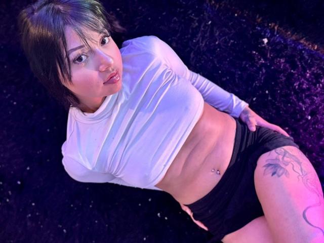 AmeliePiere with short brown hair, white long-sleeve crop top, black shorts, tattoo on right thigh, belly button piercing, lying down, purple and blue lighting.