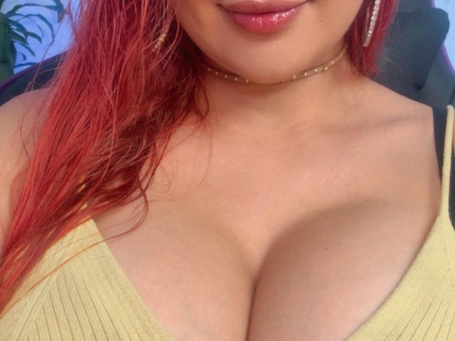 NathaPeach's red hair, shiny pink lips, yellow ribbed top with deep cleavage, showing her large, rounded, juicy tits, and a gold choker around her neck.