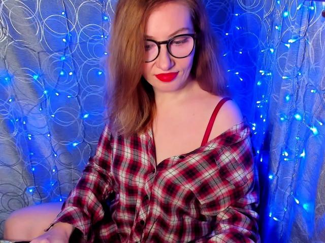 PaulinaSunny with red lipstick, glasses, and red bra strap, wearing an off-shoulder red plaid shirt, sitting against blue fairy lights, showing her tits.