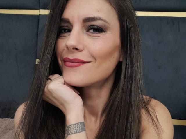 ElisBelle with long brown hair, red lipstick, and a subtle smile, resting her chin on her hand, showing a tattoo on her wrist. She's looking at the camera.