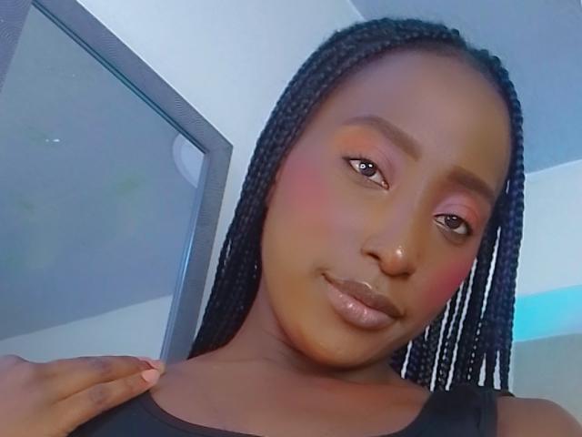 Jorinde with dark skin, long braided hair, pink blush, and glossy lips, looking at the camera with a slight smile, hand on shoulder, wearing a black top.