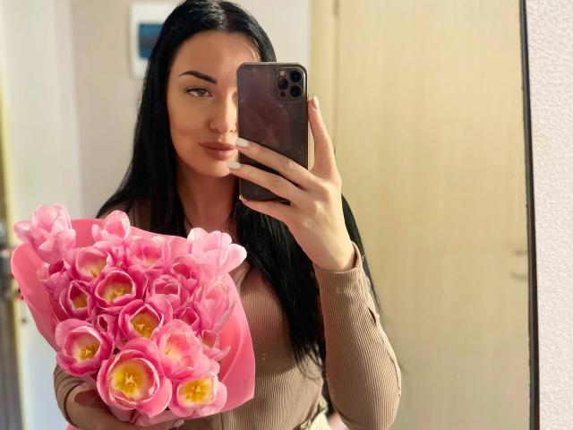 Xioni with long black hair, holding pink tulips in front of her, taking a mirror selfie with a brown phone, wearing a beige long-sleeve top, showing her tits slightly.