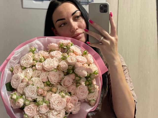 Xioni, avec des roses roses dans un emballage rose, tenant un smartphone, montrant ses ongles roses et sa bague, dans un selfie pris dans un miroir, regardant légèrement sur le côté..