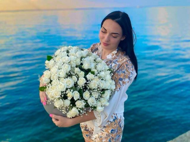 Xioni with white rose bouquet, holding it against her chest, wearing a gold-embroidered white dress, standing by blue water, long black hair, pink nails, soft smile, relaxed.