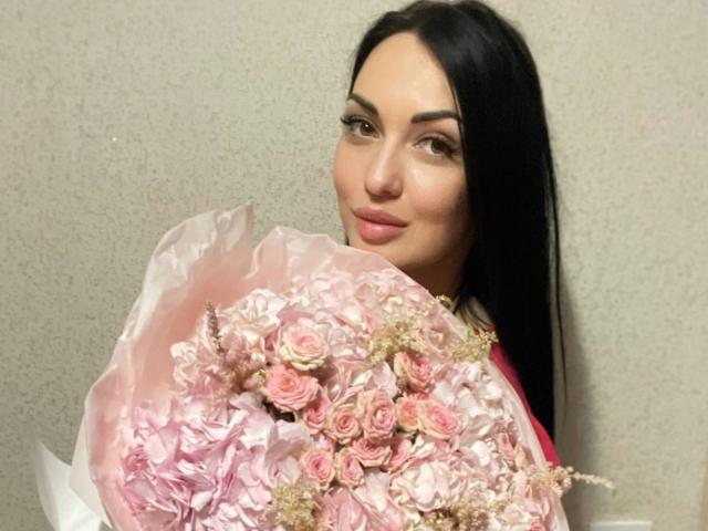 Xioni with long black hair, holding a pink rose bouquet, smiling softly. She's got a cute nose and full lips, looking sexy with her tits partly covered by flowers.