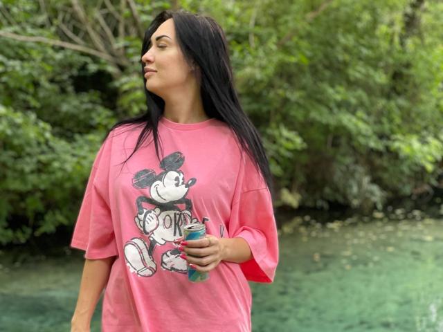 Xioni, avec des cheveux noirs, portant une chemise rose avec un dessin de singe, tenant une bouteille d'eau bleue, debout près d'une étendue d'eau verte et claire, regardant sur le côté..