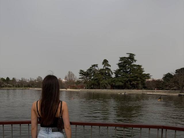 PaigePrado with long brown hair, black tank top, and blue high-waisted jeans, sitting with back to camera, looking at a calm pond with tall green trees.