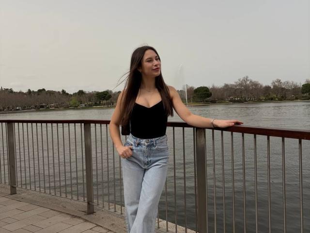 PaigePrado with long brown hair, black strapless top, and light blue high-waisted jeans, leaning on metal railing by a calm water body, hands in pockets, looking to the side.