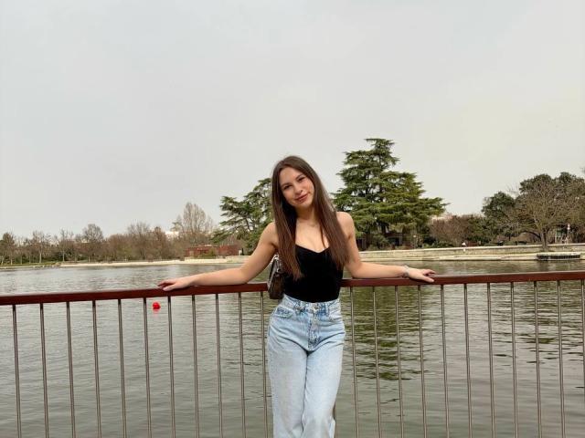 PaigePrado with long brown hair, black tank top, and high-waisted blue jeans, leaning on a metal railing by a calm, reflective waterway, hands resting on the railing.