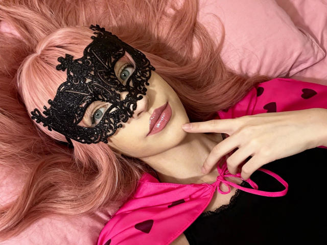 NovaVerboten-hot with pink hair, black lace mask, pink cape with heart patterns, and glossy pink lips, lying on pink pillows, touching her chin with a finger.