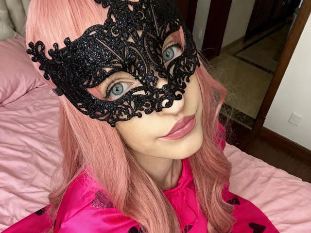 NovaVerboten-hot with pink hair, black lace mask, pink lips, and pink satin robe, showing her tits slightly, in a bedroom. She's looking at the camera with blue eyes.
