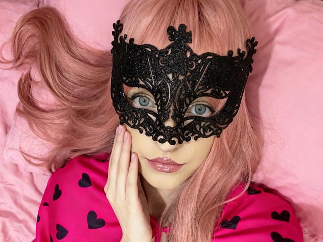 NovaVerboten-hot with pink hair, black lace mask, blue eyes, pink lipstick, and pink shirt with black hearts, touching her cheek, lying on pink pillow, looking directly at camera.