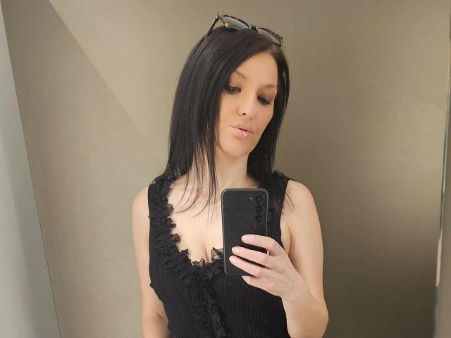 JoliDanielle with black hair, wearing a low-cut, ruffled black top, showing her tits, holding a phone, and with eyeglasses on her head, taking a mirror selfie.