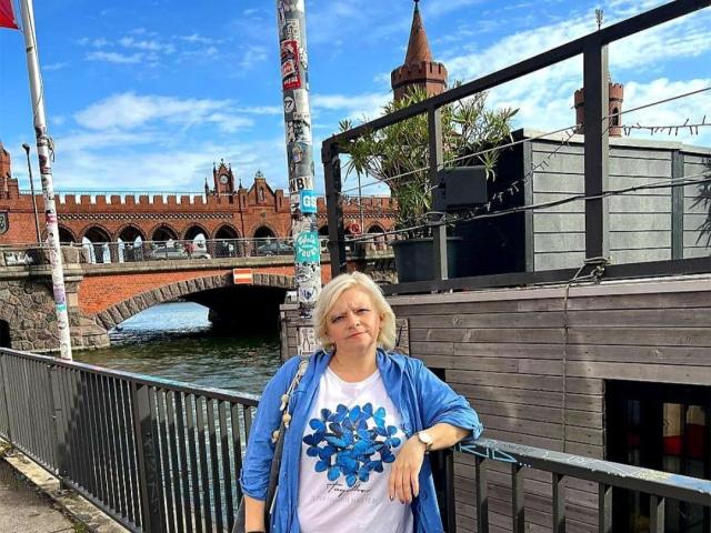 MarianneBlond with blonde hair, wearing a blue cardigan over a white shirt with blue floral pattern, standing by a metal railing, blue sky, and a brick bridge in background.