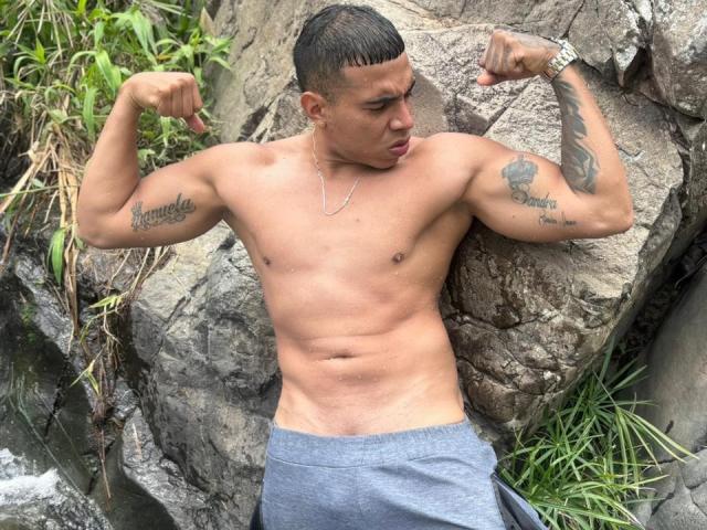 AlejandroCock flexing his muscular arms, showing tattoos on both biceps, wearing gray shorts, with a watch on his right wrist, standing against rocky background.